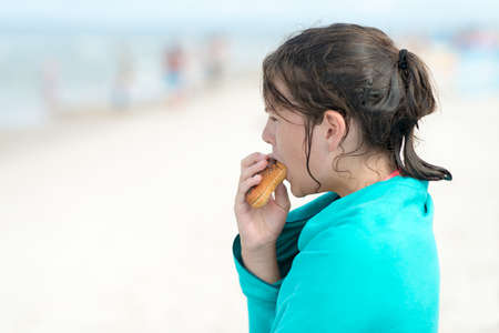 Girl wrapped in a towel after a bath in the sea, eating a sandwich on a sandy beach