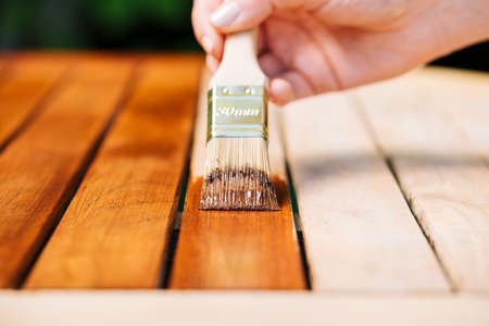 hand holding a brush applying varnish paint on a wooden garden table - painting and caring for wood with oilの写真素材