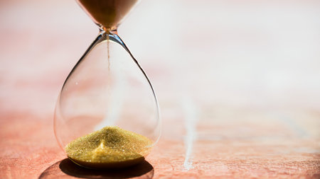 Gold Sand running through the bulbs of an hourglass measuring the passing time in a countdown to a deadline, on a light pastel reddish background with copy space - shallow depth of fieldの写真素材