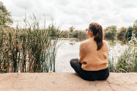 Teenage girl in a warm sweater with glasses sitting on the shore of the lake looking ahead - autumn time conceptの写真素材