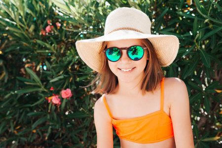 Little smiling playful girl in a straw hat and sunglasses - looking directly at the camera - against the background of a bush with flowers - holiday carefree conceptの写真素材