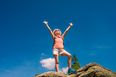 Little girl with backpack standing on top of a mountain and raising her hands upの写真素材