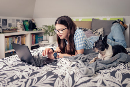 young woman with glasses and long hair is lying on a bed, working on a laptop. She is dressed in casual attire and accompanied by her black and white dog. The cozy bedroom features a patterned blanketの写真素材
