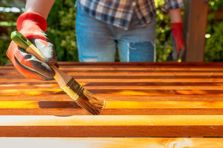 Close-up of a worker's gloved hand painting a wooden surface with a brush and stain during an outdoor DIY renovation projectの写真素材