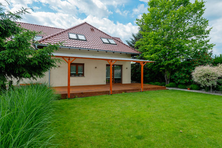 Modern house with a red tiled roof, spacious wooden deck with a pergola, and a lush green backyard surrounded by trees and landscaping on a sunny day.の写真素材