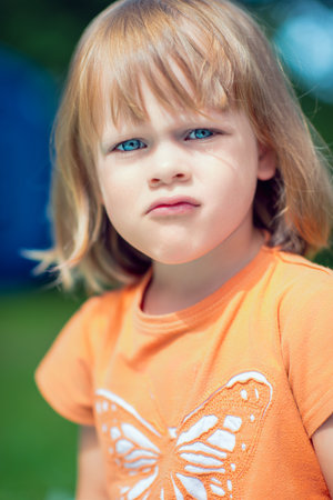 Close-up portrait of a young blonde girl with blue eyes wearing an orange butterfly t-shirt, looking...
