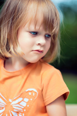 Close-up portrait of a young blonde girl with blue eyes wearing an orange butterfly t-shirt, looking thoughtful and serious outdoors in natural daylightの写真素材