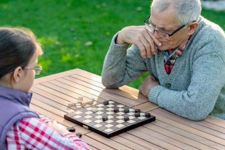 Elderly grandfather and young granddaughter playing checkers together at a wooden table in a garden, enjoying quality family time and strategic thinking outdoors in autumnの写真素材