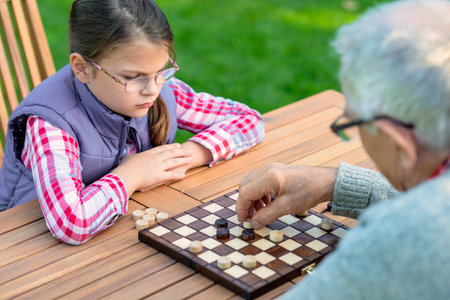 Young girl playing checkers with her grandfather at a wooden table outdoors, focusing on strategy and enjoying quality family time in a park or garden settingの写真素材