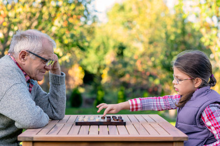 Elderly grandfather and young granddaughter playing checkers together at a wooden table in a garden, enjoying quality family time and strategic thinking outdoors in autumnの写真素材