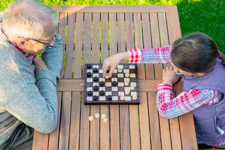 Elderly grandfather and young granddaughter playing checkers together at a wooden table in a garden, enjoying quality family time and strategic thinking outdoors in autumnの写真素材
