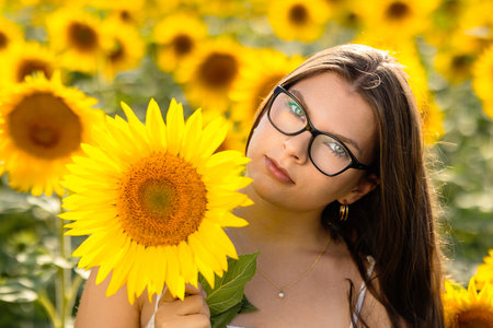 Young woman with glasses and green eyes posing with sunflower in a vibrant yellow sunflower field, beautiful brunette with gold earrings on a bright sunny summer dayの写真素材