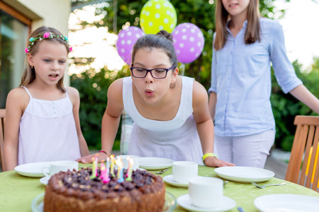 Girl with glasses blowing out birthday candles on chocolate cake while family watches, outdoor celebration with purple and green polka dot balloons, festive home party atmosphereの写真素材
