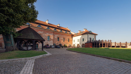 Tyniec, Poland - July 29, 2024; Panoramic view of the historic Tyniec Monastery courtyard with medieval brick and stone architecture, wooden gazebo, green lawn, parked cars, and clear blue summer skyの写真素材