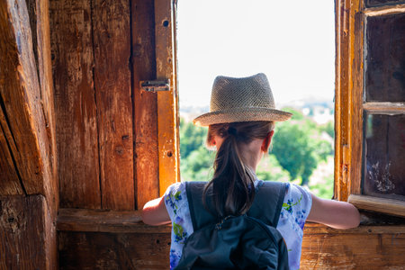 Girl in straw hat and backpack standing at rustic open window, looking out at scenic lake and forest landscape, summer adventure and travel conceptの写真素材