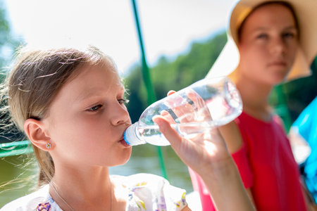 Girl drinking water from bottle outdoors in summer sunshine, staying hydrated during travel or holiday, healthy lifestyle and refreshment concept with hat and casual outfitの写真素材