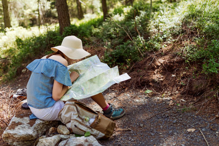 Woman hiker in wideâbrim hat studying a paper map while resting on a forest trail, outdoor navigation and route planning during a summer hike with backpack and travel gearの写真素材