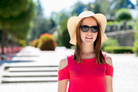 Woman in stylish wide-brim hat and pink dress standing outdoors in sunlight, summer fashion, confident portrait with blurred park background, urban lifestyleの写真素材