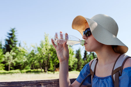 Girl in wideâbrim hat and sunglasses drinking water from a bottle while resting outdoors on a sunny day, summer hydration, travel and sunâsafety concept with copy space and natural backgroundの写真素材