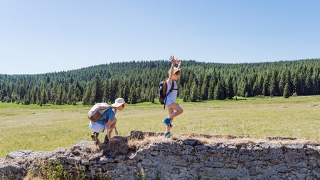Mother and daughter hikers balancing on an old stone wall in sunny mountain meadow, playful family adventure and outdoor bonding with backpacks, forest landscape and blue sky in backgroundの写真素材