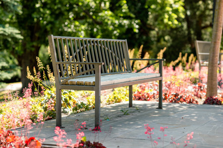 Metal garden bench among blooming flowers in sunlit park, tranquil outdoor seating area for relaxation, nature, leisure, and summer travel in public spaceの写真素材
