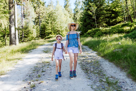 Two sisters hikers with backpacks, hats and sunglasses walking together along a sunny forest trail, summer outdoor adventure, family bonding and active lifestyle in nature sceneの写真素材