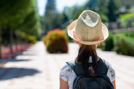 Rear view of girl in straw hat and backpack walking outdoors in sunlight, exploring city park on summer vacation, travel and adventure concept, cheerful youth lifestyle portraitの写真素材