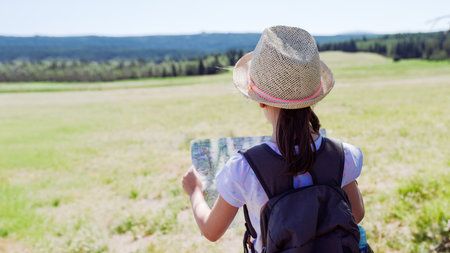 Girl hiker in straw hat and sunglasses reading a paper map in sunny mountain meadow, travel navigation and summer adventure concept with backpack, rear view and scenic landscape in backgroundの写真素材