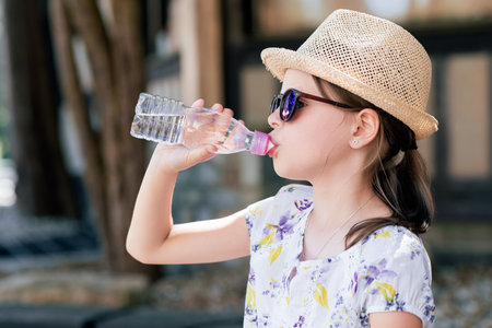 Girl in straw hat and sunglasses drinking water from a bottle outdoors, staying hydrated and refreshed on a sunny summer day during travelの写真素材