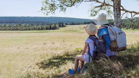 Mother and daughter hikers sitting in the shade under a tree with backpacks, looking over a sunny mountain meadow and trail, family travel, rest and bonding in nature with panoramic landscape and copy spaceの写真素材