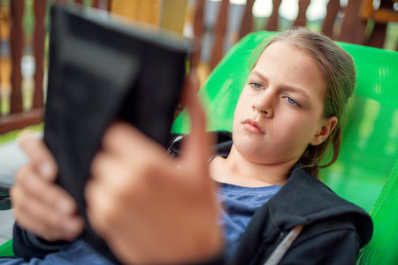 Bored teenage girl with sad pout reading on tablet while lounging in green recliner on sunny outdoor...