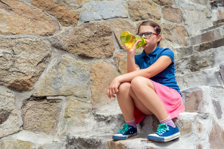 Girl in blue shirt and pink skirt sitting on old stone steps drinking water from reusable bottle during summer sightseeing breakの写真素材