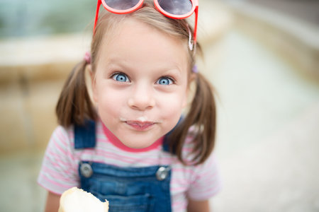 Close-up portrait of cute little girl with blue eyes and red sunglasses on head eating vanilla ice...