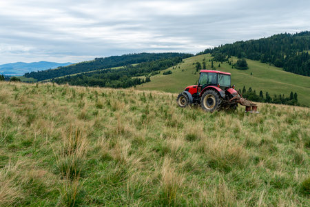 A red tractor with a harrow on a grassy hillside, amidst forested mountains and green valleys under an overcast sky, mowing thistlesの写真素材
