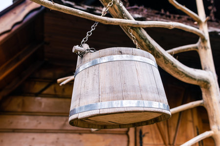 Hanging Wooden Well Bucket with Metal Bands and Chain in Front of Rustic Timber House, Vintage Rural Decoration Detailの写真素材