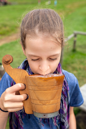 A delighted little girl with her eyes closed tries a traditional, thick drink made from sheep's whey, made in Podhale from a wooden ladle and a purple scarf, on a bench in a meadowの写真素材