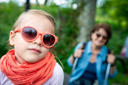 Cute Toddler Girl in Orange Sunglasses and Lace Scarf Portrait with Blurred Hiking Grandma on Forest Trailの写真素材