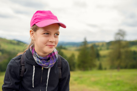 Pensive Teenage Girl in Pink Cap and Plaid Scarf Gazes over Green Rolling Hills, Backpack On, Cloudy Countryside Portraitの写真素材