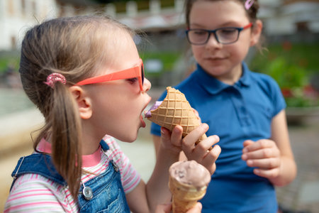Two sisters sharing waffle cone ice cream together in sunny city park, close-up of kids hands and faces, sweet summer dessert momentの写真素材