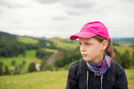 Thoughtful Teen Girl in Pink Cap, Plaid Scarf, and Backpack Looks Sideways over Lush Green Hills and Cloudy Skyの写真素材