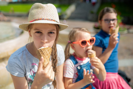 Happy group of children eating ice cream cones together by city park fountain on sunny summer day, wearing colorful casual clothes and hatsの写真素材