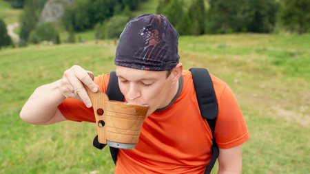 An adult hiker in an orange shirt drinks from a rustic wooden cup outdoors in a green meadow during a break in his hikeの写真素材