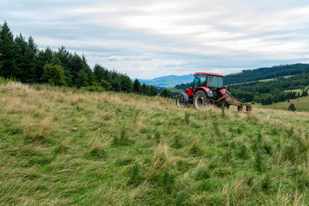 A red farm tractor with an attachment mowing thistles on a grassy hillside overlooking green pastures and distant mountains under an overcast skyの写真素材