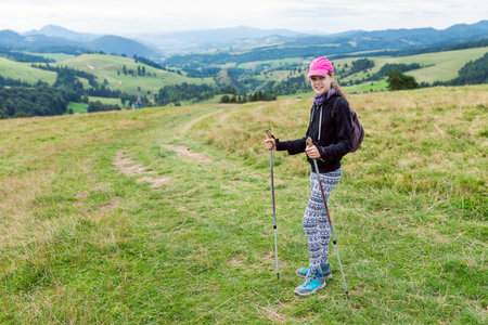 Young Female Hiker with Pink Cap, Patterned Leggings, Trekking Poles and Backpack on Grassy Hill overlooking Green Mountain Valley Panoramaの写真素材
