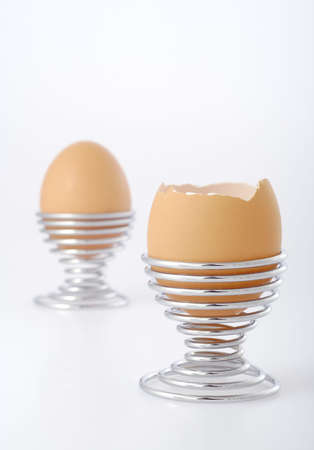 Two eggs in wire eggcups on bright background. Focus on front egg with partially removed eggshell....