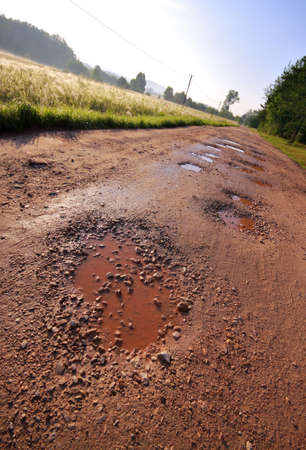 Local red clay road with wet holes surrounded by field and forest. Oblique shoot.の写真素材