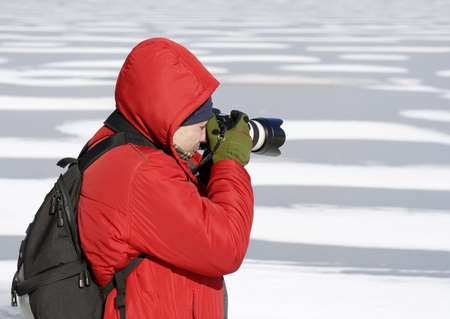 Photographer looking through camera with red jacket and backpack on unfocused freezed lake backgroundの写真素材