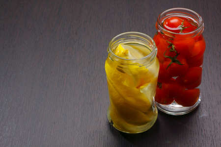 Two opened jars with lemon wedges and cocktail tomatoes on dark board. Copy space on left.