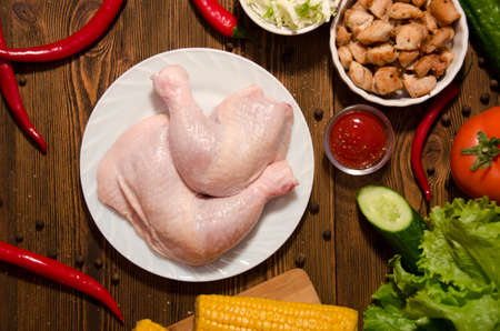 raw chicken legs on a white plate on a wooden background with vegetables and pepper top viewの写真素材