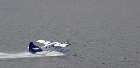 A blue and white float plane takes off from a body of water  spray from the pontoon is seen as it...